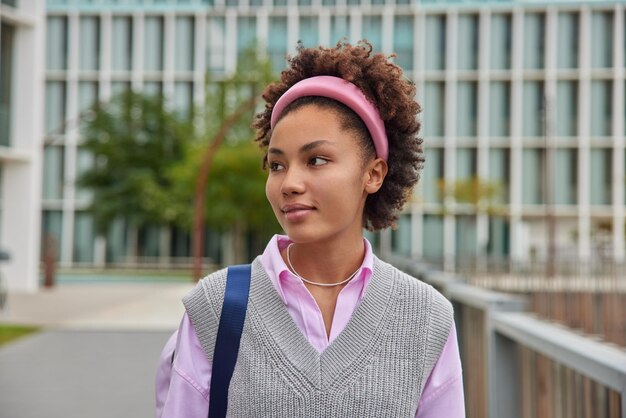 Outdoor shot of pensive curly haired woman student returns from lecures wears headband and casual neat clothes strolls outdoors poses against blurred urban background People and lifestyle concept