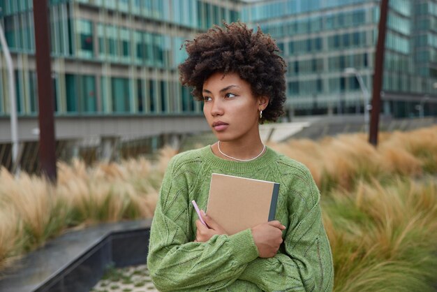 物思いにふける縮れ毛の若い美しい女性の屋外ショットは、カジュアルな緑のジャンパーに身を包んだ目をそらしますノートと鉛筆を保持しますどこかに焦点を当てたモダンな都市の建物の背景の近くに立っています