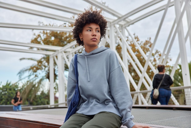 Free photo outdoor shot of pensive curly haired young woman concentrated into distance dressed in hoodie and trousers thinks about something rests after excursion in city feels tired explores new places