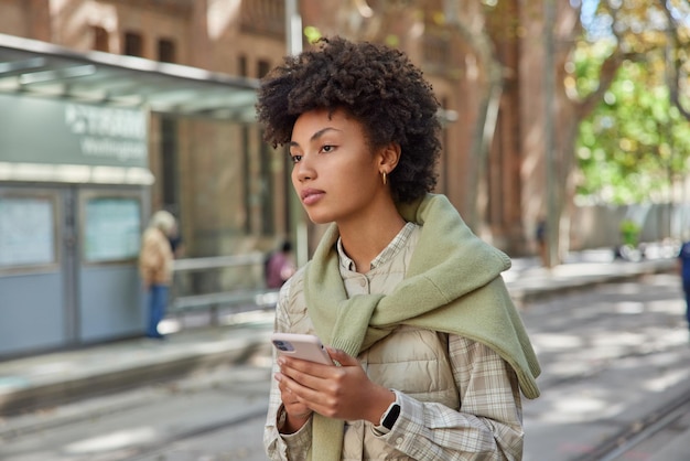 Outdoor shot of pensive curly haired young woman holds mobile phone chats online tries to find way in unknown city wears casual outfit