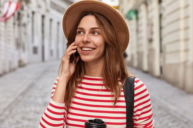 Outdoor shot of pleased Caucasian traveler talks on smart phone in roaming, enjoys cheap tariffs for communication, stands on street