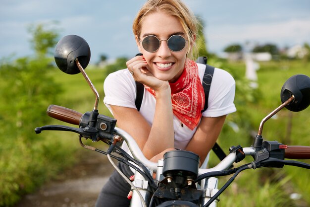 Outdoor shot of positive active female driver sits on fast motorbike, wears fashionable clothing, has break after biker`s competition in countryside. People, motorcycling and lifestyle concept