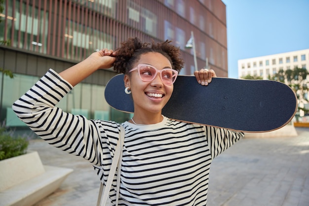 Outdoor shot of pretty cheerful millennial girl dressed in casual striped jumper carries longboard leads active lifestyle smiles gladfully poses against urban background Positive female skater