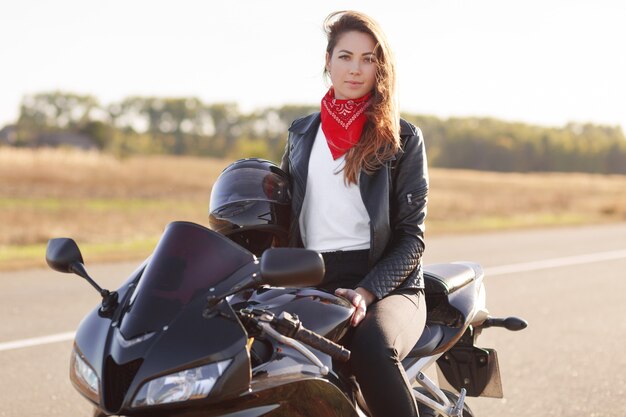 Outdoor shot of pretty woman biker wears red banadana and leahter jacket