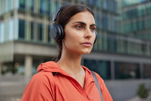 Outdoor shot of serious brunette young woman leads sporty lifestyle listens music via headphones enjoys playlist exercises in city dressed in anorak poses at street takes break after workout