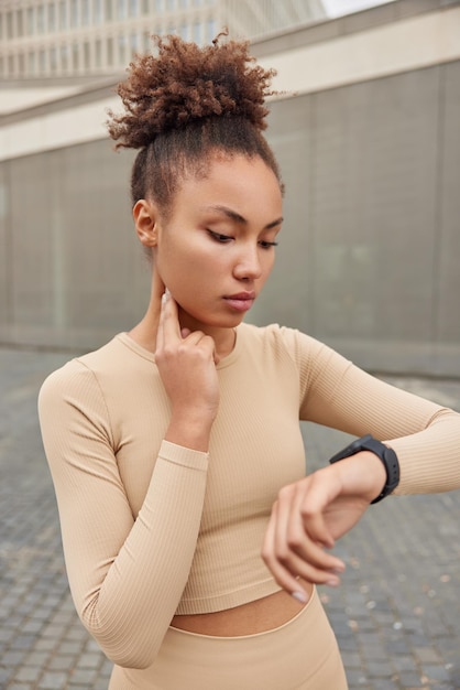 Outdoor shot of serious woman with curly hair gathered in bun checks heart rate focused at smartwatch monitors pulse dressed in activewear leads healthy lifestyle goes in for sport regularly