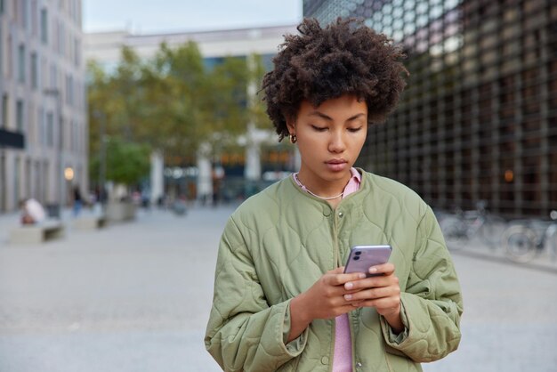 Outdoor shot of serious woman with curly hair uses smartphone types sms focused at cellular screen wears jacket strolls at street during daytime People modern technologies and lifestyle concept