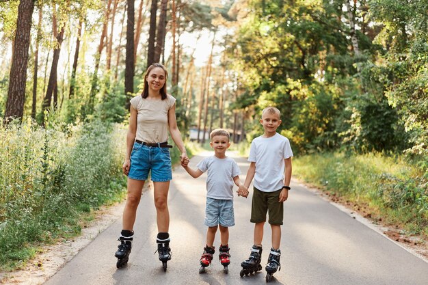 Открытый снимок улыбающейся привлекательной женщины с ее маленькими сыновьями, стоящими на дороге в летнем парке и держащимися за руки, семьями, катающимися на роликах, весело, активным времяпрепровождением.