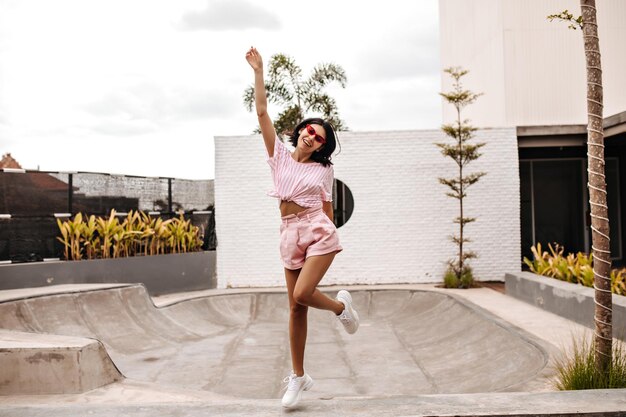 Открытый снимок стильной дамы, прыгающей с улыбкой Вид загорелой девушки в розовом летнем наряде в полный рост