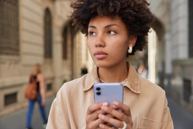 사려 깊은 곱슬머리의 아름다운 여성이 휴대전화를 탐색하는 야외 사진은 여행 중에 내비게이션 앱을 사용하여 도시 거리를 걷는 동안 콘텐츠 공유 멀티미디어를 온라인에 집중적으로 보냅니다.