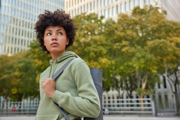 Outdoor shot of thoughtful sportswoman in hoodie carries karemat poses against city background with green trees concentrated into distance goes in for sport regularly. Yoga practice and recreation