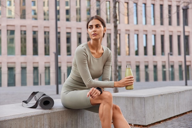 Outdoor shot of thoughtful sporty young woman dressed in activewear takes break after sport training feels thirsty drinks water from sport bottle sits and looks away pensively uses fitness mat