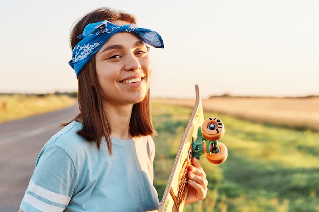 Outdoor shot of young adult winsome woman wearing blue casual style t shirt and hairband ,standing with longboard in hands, looking at camera with charming smile.