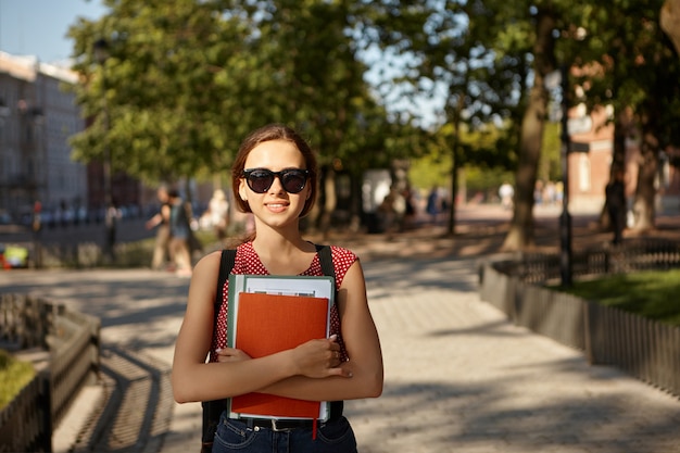 Outdoor summer image of adorable cute Caucasian female student wearing stylish shades, backpack, dotted top and jeans commuting to college by foot, carrying copybooks, smiling, enjoying nice weather