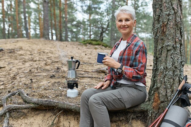 Outdoor summer image of cheerful middle aged woman in activewear relaxing under tree with camping gear and kettle on gas burner, holding mug, enjoying fresh tea, having rest while hiking alone