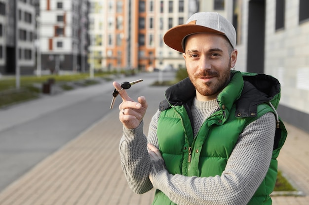 Foto di estate all'aperto di felicissimo bel giovane barbuto con espressione facciale allegra e sicura, sorridente, torcendo i tasti intorno al dito indice, orgoglioso della nuova auto, appartamento, in piedi all'aperto