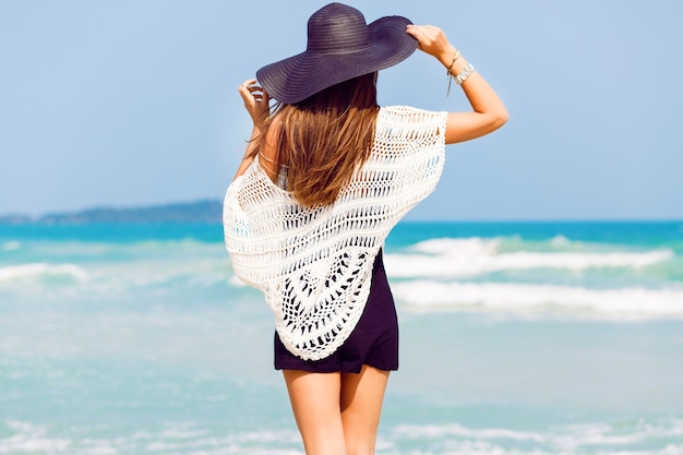 Открытый летний портрет молодой красивой женщины, смотрящей на океан на тропическом пляже, наслаждающейся ее свободой и свежим воздухом, в стильной шляпе и одежде