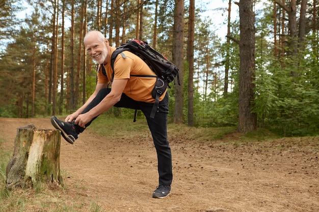 Outdoor summer shot of healthy fit elderly male with backpack posing in forest with foot on stub, tying shoelaces on sneakers, getting ready for long climb, hiking with happy smile