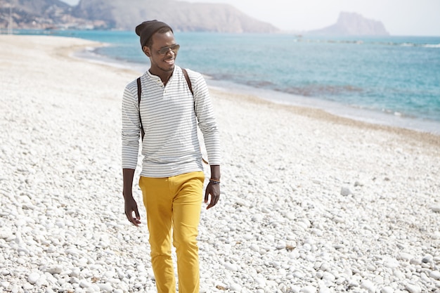 Outdoor summer shot of young African American tourist in hat and stylish sunglasses, carrying backpack on his shoulders, walking along pebble beach, looking happy and carefree, enjoying seascape