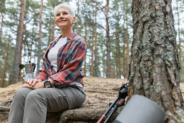 Outdoor summer view of attractive adventurous middle aged female sitting by tree, boiling water for tea in kettle, having joyful look, admiring beautiful nature, birds singing, smiling happily