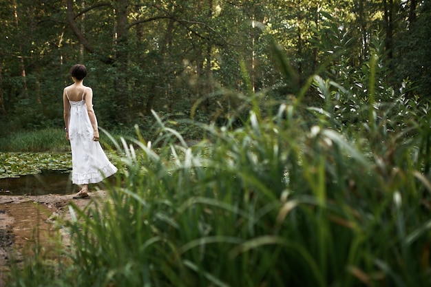週末に一人で野生の自然の中でリラックスし、前景に新鮮な緑の草と背景の池のそばに立って、長い白いドレスを着てロマンチックな愛らしい若い女性の屋外の夏のイメージ