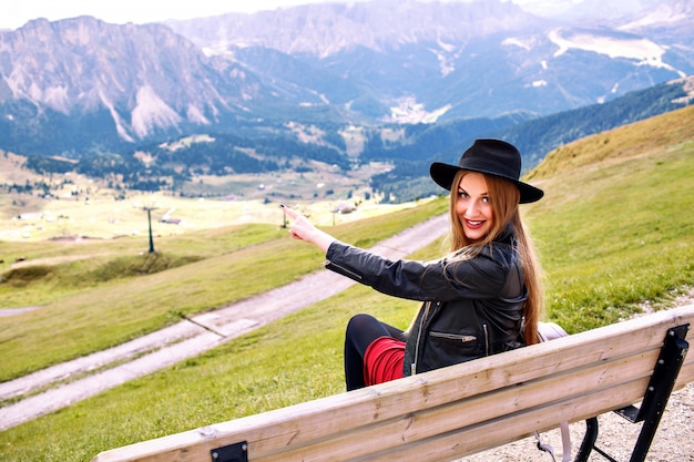 Outdoor travel image of stylish woman sitting in the bench at mountains resort, showing by her hand on amazing view to Alp mountains, luxury trip.