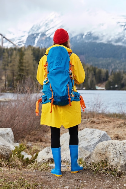 Outdoor vertical shot of female tourist admires turquoise water from lake, stands near rocks, looks at snow mountains, breathes fresh air, wears red hat