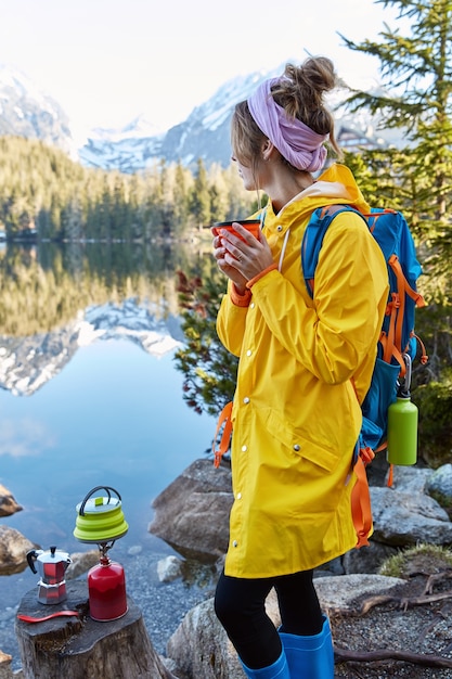 Outdoor vertical shot of pensive female hicker holds hot beverage in teacup, makes drink on special tourist equipment