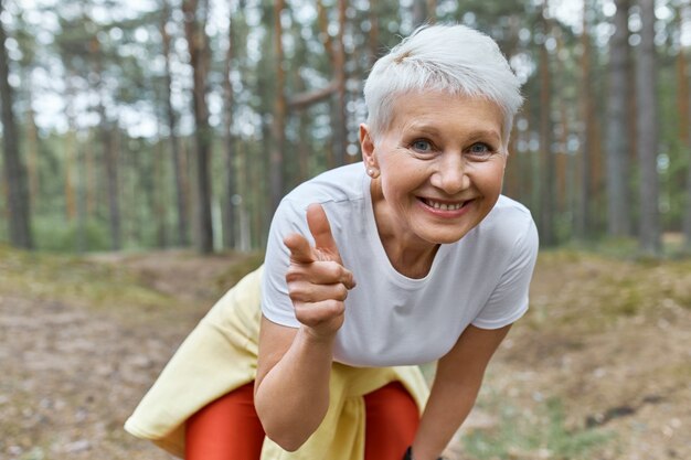 Outdoor view of energetic joyful female pensioner in sports clothes leaning forward, smiling and pointing fore finger at front