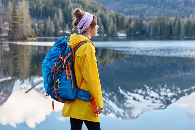 Outdoor view of thoughtful restful woman stands near calm lake with mountain reflection