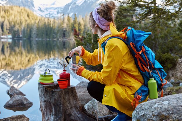 Outdoor view of young woman uses tourist equipment for making coffee, has portable gas stove on stump