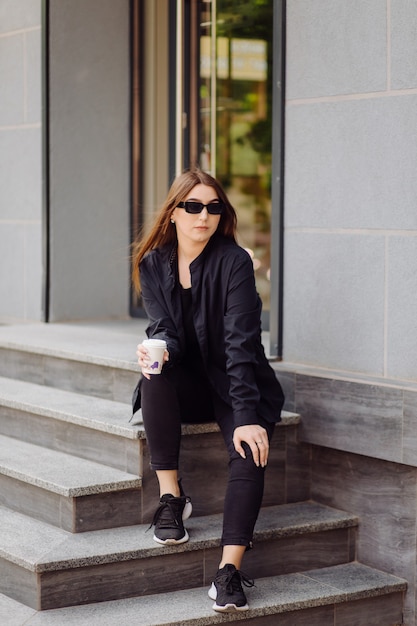 Outdoors lifestyle portrait of stunning brunette girl. Drinking coffee and walking on the city street.