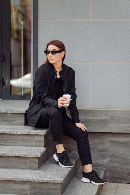 Outdoors lifestyle portrait of stunning brunette girl. Drinking coffee and walking on the city street.