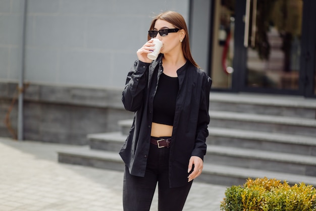 Outdoors lifestyle portrait of stunning brunette girl. Drinking coffee and walking on the city street.