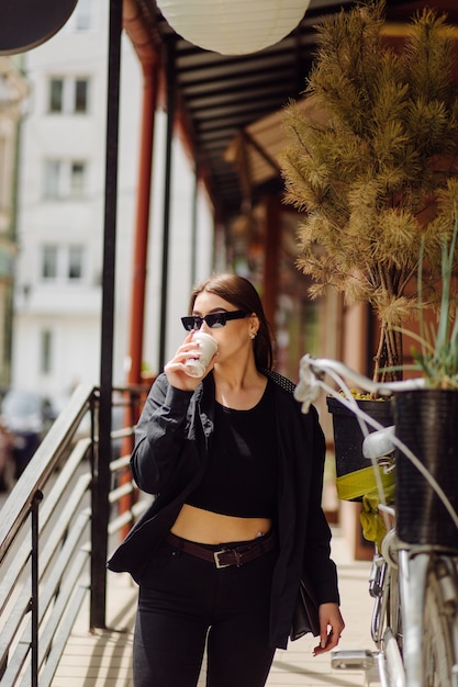 見事なブルネットの少女のアウトドアライフスタイルの肖像画。コーヒーを飲み、街の通りを歩きます。