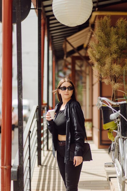 Outdoors lifestyle portrait of stunning brunette girl. Drinking coffee and walking on the city street.