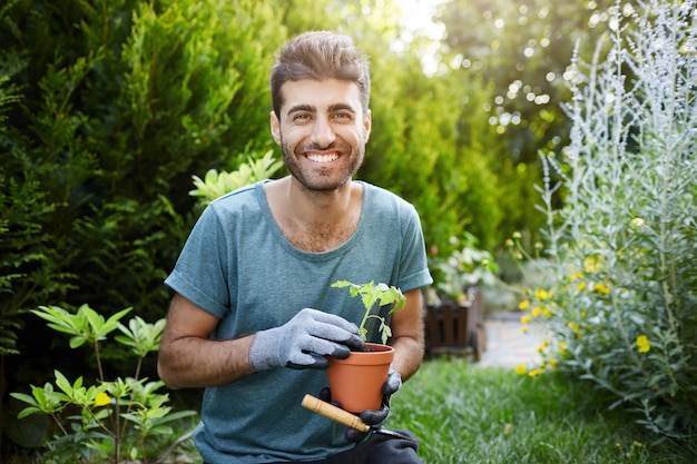 Outdoors portrait of young good-looking caucasian bearded man in blue shirt and gloves smiling in camera, holding pot with flower in hands working in garden.