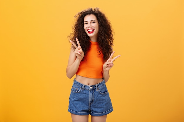 Outgoing friendly-looking attractive young female with curly haircut in cropped top and shorts showing peace or victory signs with both hands and smiling cheerfully having fun over orange background.