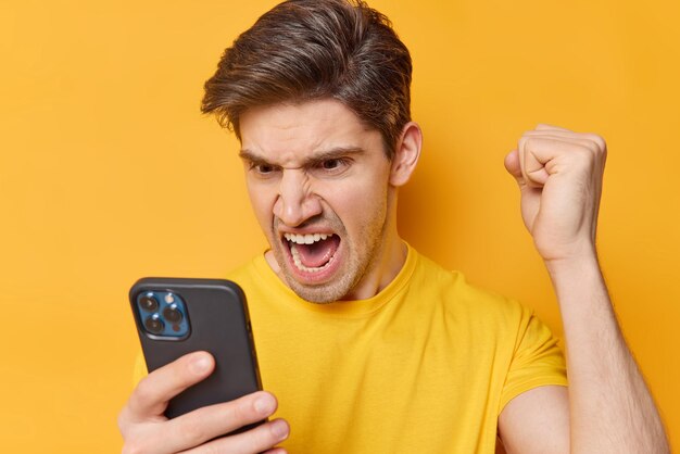 Outraged angry adult man screams loudly focused at smartphone screen clenches fist has argument with something during distance call dressed in casual t shirt isolated over yellow background