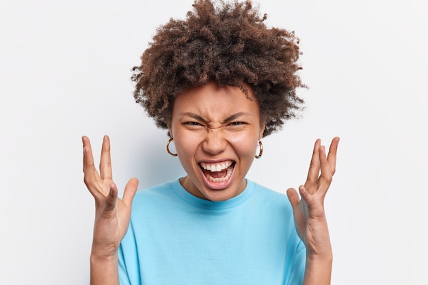 Outraged annoyed young curly African American gestures angrily screams from anger and disappointment shakes hands looses temper yells upset isolated over white wall. Negative emotions.