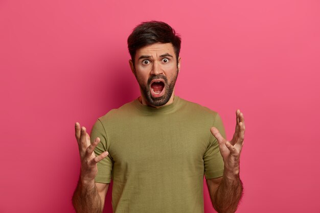 Outraged bearded man gestures angrily, exclaims loudly, keeps mouth opened, has bothered displeased expression, agrues with someone, grimaces hateful , poses against pink wall