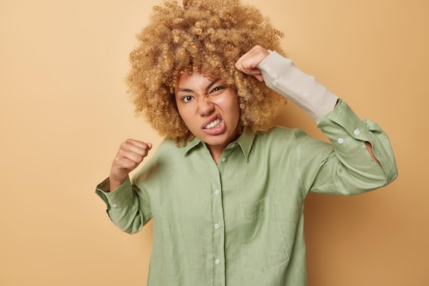 Outraged curly haired young woman clenches fists and looks angrily has broken arm wears bandage dressed in casual green shirt isolated over brown background People aggression rage and anger