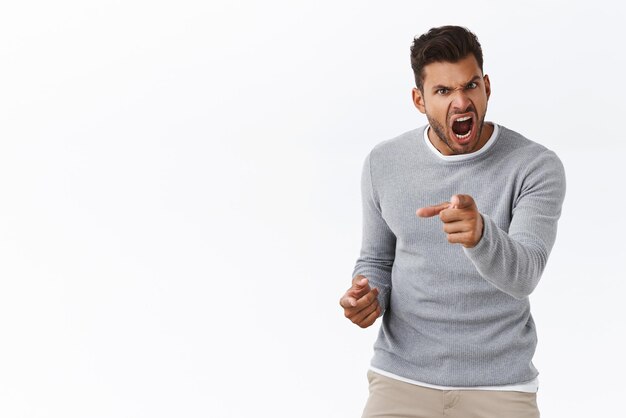 Outraged handsome man with bad temper losing control over emotions distressed pointing camera and shouting accusations blame someone in anger and furious feelings standing white background