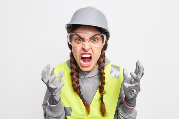 Outraged irritated female construction worker gestures angrily screams loudly being annoyed with partners who made big failure or mistake wears protective hardhat gloves safety glasses uniform