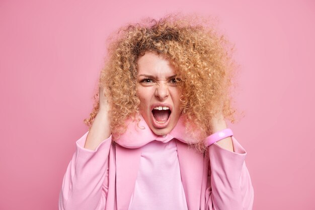 Outraged mad young woman with curly hair screams loudly being very angry keeps mouth opened wears travel pillow around neck formal clothes isolated over pink wall. Human emotions concept