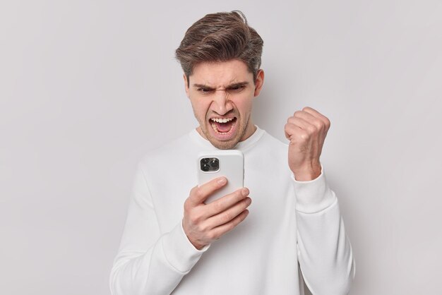 Возмущенный мужчина сердито смотрит на смартфон, получает раздражающее сообщение, сжимает кулак и громко восклицает, носит случайный джемпер, изолированный на белом фоне, спорит с телефоном, выражает негативные эмоции