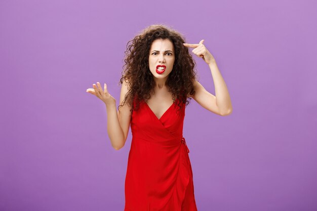 Outraged and pissed wife critisize dumb idea, outraged and fed up with stupid husband actions rolling finger near temple gesturing with raised palm, being angry and questioned over purple background.