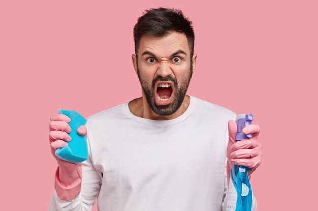 Outraged unshaven man carries blue mop and detergent, dressed in white clothes, feels angry with wife who makes him cleaning house