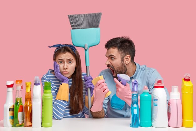Outraged unshaven man shouts at wife, complains of much work, carries spray and rag, displeased woman wears headband, carries broom