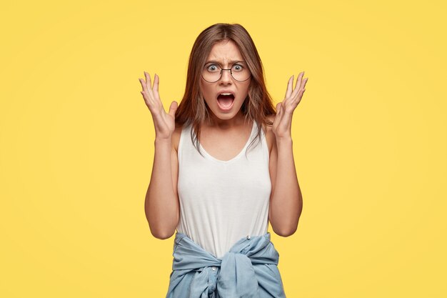 Outraged young brunette with glasses posing against the yellow wall